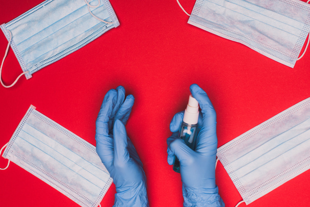 Top view of doctor in latex gloves holding hand sanitizer near medical masks on red background - Photo, Image