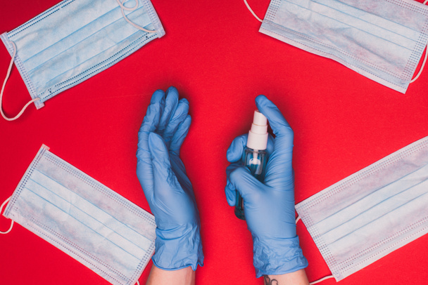 Vista superior del médico sosteniendo el desinfectante de manos cerca de máscaras médicas sobre fondo rojo
 - Foto, Imagen