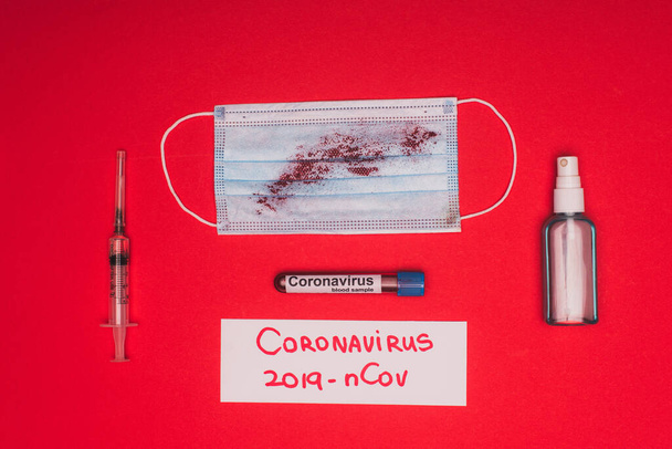 Kanlı tıbbi maskenin üst görüntüsü, şırınganın yanındaki el dezenfektanı kan örneğiyle test tüpü ve kırmızı arka planda Coronavirus 2019-nCov harfleri. - Fotoğraf, Görsel