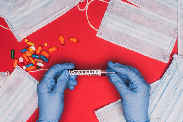 Top view of doctor holding test tube with blood sample and coronavirus lettering near pills and medical masks on red background - Photo, Image