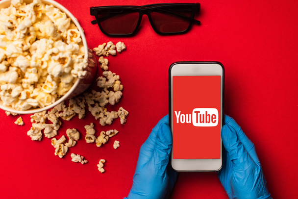 KYIV, UKRAINE - MARCH 26, 2020: Top view of person holding smartphone with youtube app near popcorn and sunglasses on red background - Фото, изображение