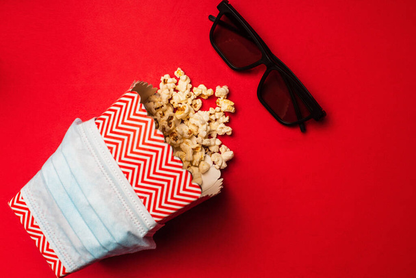 Top view of medical mask on bucket with popcorn near sunglasses on red background - 写真・画像