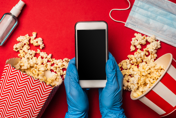 Vista dall'alto di persona che tiene smartphone con schermo vuoto vicino alla maschera medica e popcorn sulla superficie rossa
 - Foto, immagini