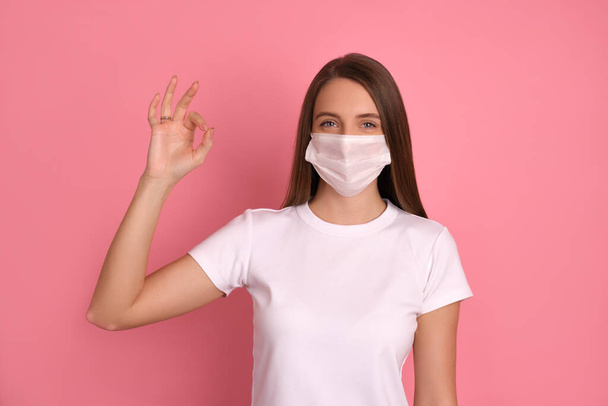 Happy brunette girl wears a white medical mask to protect her from Corona virus, cares for her health and safety, has a beautiful smile, sticks to self-isolation, shows ok ring-gesture to be positive. Covid-19 - Фото, изображение