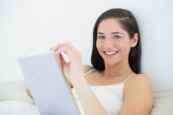 Close-up portrait of a smiling woman with book in bed - Foto, Bild
