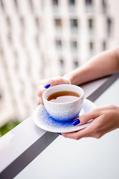 Weibliche Hände halten eine Tasse Tee auf dem Balkon. Maniküre mit lebendiger blauer Farbe Nagellack - Foto, Bild