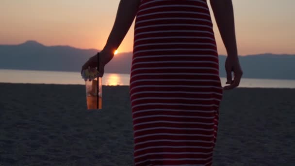Close-up de menina sexy em vestido de verão listrado vermelho segurando mojito fresco saboroso e girando na praia do mar em fundo incrível pôr do sol. Festa de praia, férias e férias de verão. Relaxar conceito
 - Filmagem, Vídeo