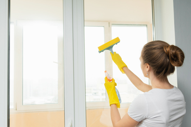 かわいい大人の女の子はスクレーパーで窓を洗う。春の掃除。清掃会社の従業員が家の中のプラスチック窓を拭く. - 写真・画像