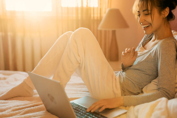 Joyful woman laughting and talking on video call on laptop computer in bed at home, sunlight in bedroom. Positive girl chatting with friends in social networks on laptop device using wireless internet - Photo, Image