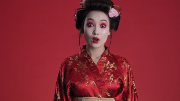 A surprised young geisha woman in traditional japanese kimono is rejoicing standing isolated over red background - Felvétel, videó