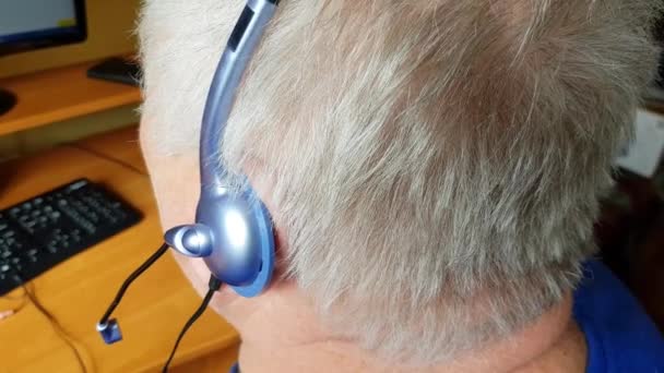 Un anciano canoso con auriculares utiliza una computadora para aprender a distancia, escucha música y se comunica a través de Skype. Estilo de vida de jubilación activo, vista desde atrás
 - Metraje, vídeo