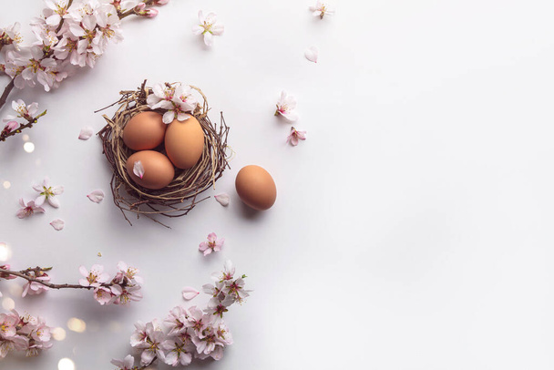 Пасхальные яйца в гнезде и розовые цветущие ветви сакуры на белом фоне с боке, свет. С праздником Пасхи, вид сверху, плоская постель - Фото, изображение
