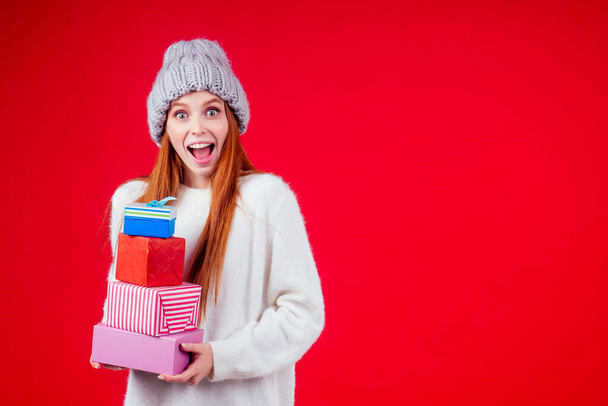 redhaired ginger woman wear stylish wool hat and white sweater looking at camera and holding three boxes witg gifts copyspase in studio red background - Photo, Image
