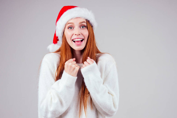 beautiful redhaired ginger woman wearing white knitted sweater and santa claus hat finger cross making wishes in studio background - Fotografie, Obrázek