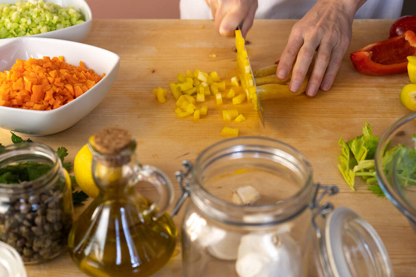 Moments of daily life in the Mediterranean cookery: low angle view of a young female cook cutting a yellow pepper with various vegetables around on her light wooden work table with bokeh effect - Photo, Image