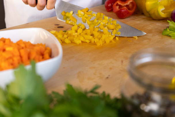 Στιγμές της καθημερινής ζωής στη μεσογειακή κουζίνα: άποψη χαμηλής γωνίας μιας νεαρής μαγείρισσας που κόβει ένα κίτρινο πιπέρι με διάφορα λαχανικά γύρω από το ελαφρύ ξύλινο τραπέζι εργασίας της με bokeh αποτέλεσμα - Φωτογραφία, εικόνα