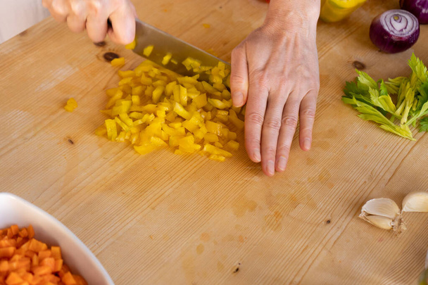 Momente des Alltags in der mediterranen Küche: Tiefblick einer jungen Köchin, die auf ihrem hellen Holzarbeitstisch mit Bokeh-Effekt eine gelbe Paprika mit verschiedenen Gemüsesorten schneidet. - Foto, Bild
