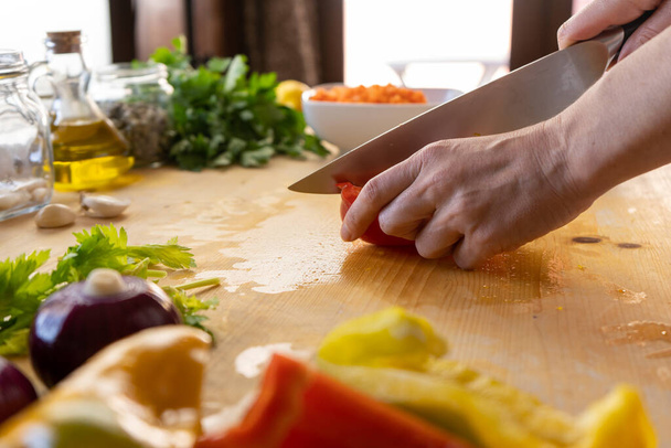 Στιγμές της καθημερινής ζωής στη μεσογειακή κουζίνα: άποψη χαμηλής γωνίας μιας νεαρής μάγειρας που κόβει μια κόκκινη πιπεριά με διάφορα λαχανικά γύρω από το ελαφρύ ξύλινο τραπέζι εργασίας της με bokeh αποτέλεσμα - Φωτογραφία, εικόνα