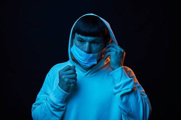 Porträt junger Modemann in weißem Kapuzenpulli im Kapuzenmantel in Medizinmaske in farbigen Lichtern auf schwarzem Hintergrund - Foto, Bild