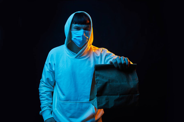 young delivery man in white clothes in rubber mask with a yellow backpack on a black background with paper bag - Zdjęcie, obraz