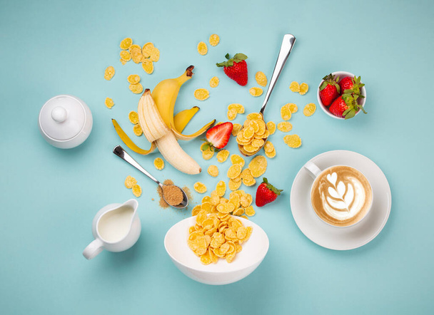 朝の迅速な健康的な朝食を準備:穀物を追加,白いボウルに果物.グラノーラと朝食を作るための成分,ミルク,青の背景に新しく作られたカプチーノ,トップビューセット - 写真・画像