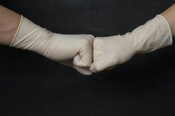 Powitanie pięści do pięści w medycznych gumowych rękawiczkach. Ręce w białe medyczne rękawice gumowe lateksowe na czarnym tle. Rękawice ochronne jednorazowe chronią przed wirusami, koronawirusami, niebezpiecznymi bakteriami. - Zdjęcie, obraz