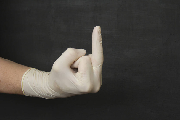 Une main dans un gant en latex médical blanc jetable montre le majeur sur un fond noir, Va te faire foutre. Gants jetables protecteurs protègent contre les virus, coronavirus, bactéries dangereuses
. - Photo, image