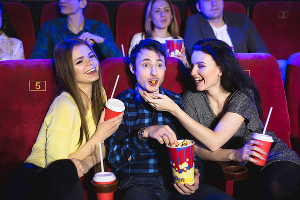 2人の若い女の子と男が映画の中でコメディを見ています。映画館で映画を見ている若い友達。ポップコーンとドリンクを持つ劇場の人々のグループ. - 写真・画像