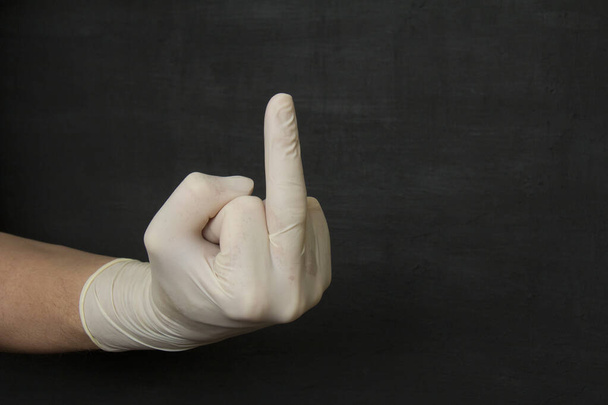 Una mano in un guanto di lattice di gomma monouso medico bianco mostra il dito medio su uno sfondo nero, Vaffanculo. Guanti protettivi monouso proteggono contro virus, coronavirus, batteri pericolosi
. - Foto, immagini