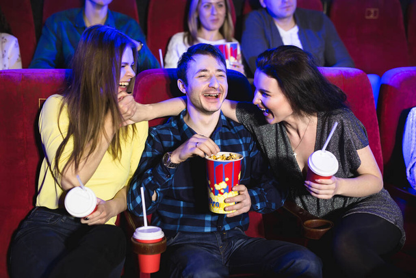Two young girls and a guy watching a comedy in a cinema. Young friends watching movie in cinema. Group of people in theater with popcorn and drinks. - Photo, Image