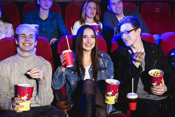 Two young guys and a girl watching a Comedy in a movie theater. Young friends watch movies in the cinema. Group of people in the theater with popcorn and drinks. - Photo, Image