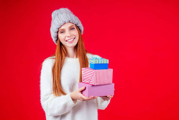 redhaired ginger woman wear stylish wool hat and white sweater looking at camera and holding three boxes witg gifts copyspase in studio red background - Fotó, kép