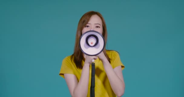 Speaking  Loud noise announce of young asian woman with megaphone in yellow T-shirt smiling emotion on blue background isolated studio shot  - Materiał filmowy, wideo