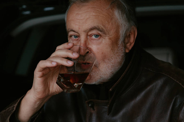 elderly man with a gray beard in a leather jacket drinks whiskey from a glass glass against the background of an open trunk of a passenger ca - Valokuva, kuva