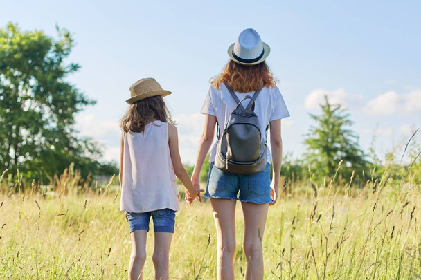 Summer vacation, children two girls sisters walking together holding hands, blue sky meadow background. Kids in hats with backpacks, back view - Foto, afbeelding