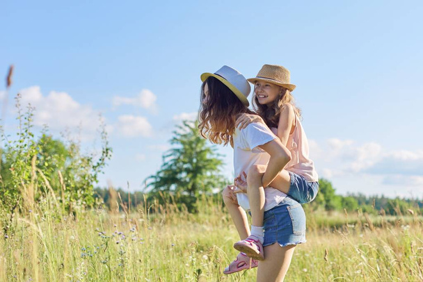Happy children two girls sisters teenager and younger laughing and having fun in meadow, blue sky, summer nature. Active healthy lifestyle, friendly family, happy childhood, copy space - Photo, image