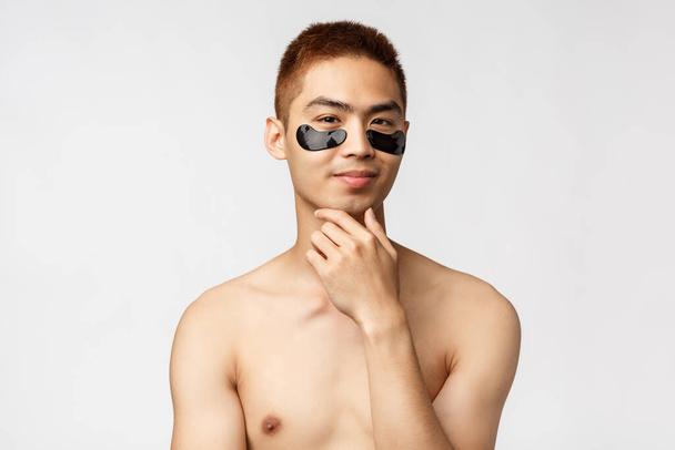 Beauty, people and lifestyle concept. Thoughtful enthusiastic, handsome naked asian man wearing eye-patches and smiling, have an idea, thinking, consider apply skincare mask, white background - 写真・画像