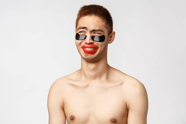 Beauty, people and leisure concept. Funny and silly asian naked boyfriend, laughing and looking away, girlfriend making fun of him applying eye-patches and lip-patch, white background - Photo, Image