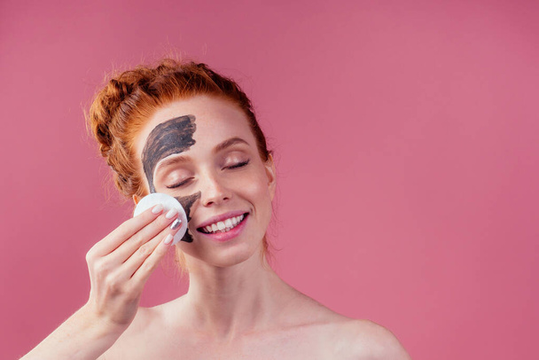 redhaired ginger teenager girl remove with cotton black clay mask on her pretty face on pink studio background - Zdjęcie, obraz