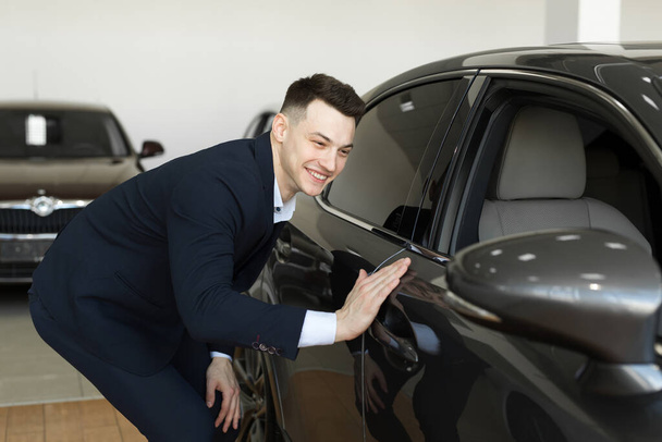 Knappe jonge zakenman in klassiek blauw pak lacht tijdens het onderzoek van de auto in een autoshow. - Foto, afbeelding