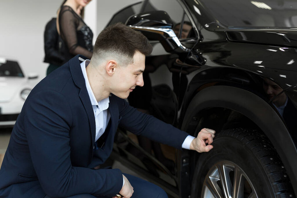 Homme élégant attrayant examinant les roues d'une nouvelle voiture en vente chez le concessionnaire. Charmant chauffeur masculin choisir une nouvelle voiture à acheter
 - Photo, image