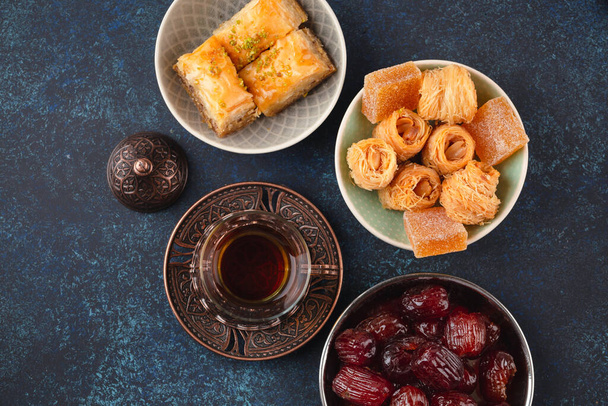 Традиционный арабский свежий горячий чай со сладостями, пахлавой, финиками, локумом сверху. Рамадан закуски с турецкими деликатесами и арабской выпечки на голубом деревенском фоне вид сверху
. - Фото, изображение