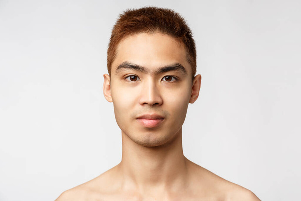 Kauneus, ihonhoito ja miesten terveyden käsite. Komea nuori aasialainen mies, jolla ei ole vikoja, täydellinen ihosairaus ilman aknea, hymyilevä innostunut, seisova valkoinen tausta
 - Valokuva, kuva