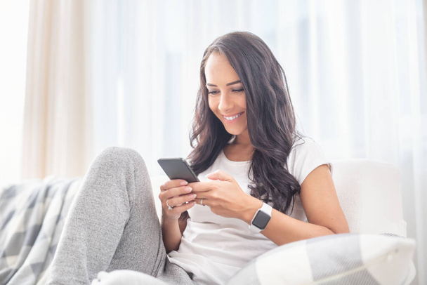 Smiling young female sitting on a couch in a living room smiling into the phone spending time online during a lockdown.  - Φωτογραφία, εικόνα