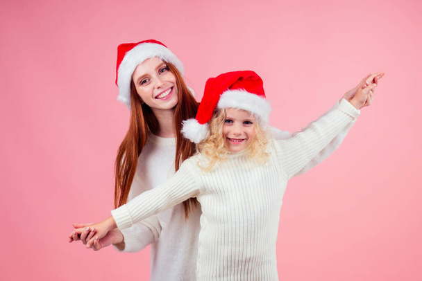 happy redhead ginger woman and cute little blonde girl wearing santa claus hat and having fun together hands like airplane wings in studio pink background. xmas journey concept - Fotoğraf, Görsel