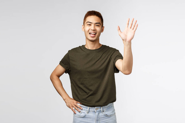 Mensen, emoties en levensstijl concept. Portret van vriendelijke vrolijke aziatische man die gedag zegt, welkomstpersoon als zwaaiende hand, informele begroeting, hallo leuk om je te ontmoeten, staande grijze achtergrond - Foto, afbeelding