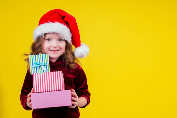 amazed and surprised little girl wear santa claus hat looking at camera and holding three boxes witg gifts copyspase in studio yellow background - Photo, Image