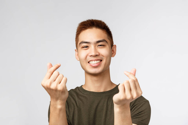 Mensen, verschillende uitdrukkingen en levensstijl concept. Schattige aziatische kerel met stijlvol kapsel, tonen koreaanse vinger harten en glimlachen mooi, uiten sympathie of als, staan grijze achtergrond - Foto, afbeelding