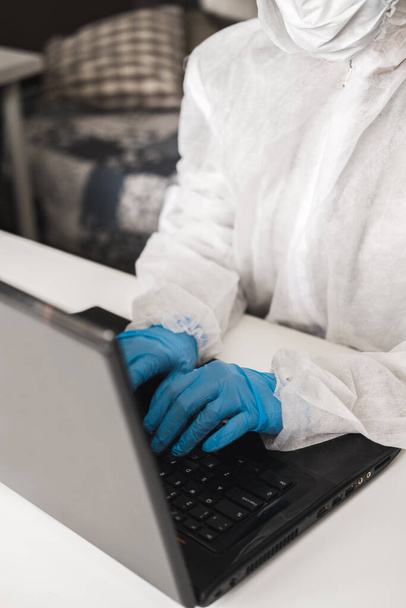 Person in protective white translucent suit, blue rubber gloves, medical mask sits at table at home and works or studies on laptop during quarantine. Remote work during coronavirus pandemic. - Photo, image