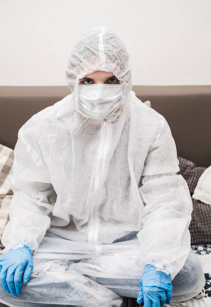 Person in protective white translucent suit, blue rubber gloves, medical mask sits at table at home and works or studies on laptop during quarantine. Remote work during coronavirus pandemic. - Valokuva, kuva